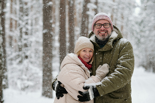Cheerful mature couple in winterwear looking at camera during snowfall in forest covered with snow while man embracing his wife