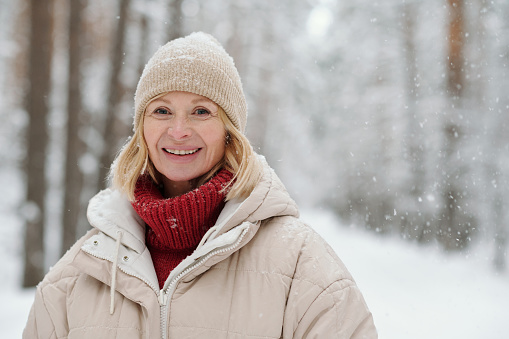 Happy mature blond woman in woolen knitted beanie, red sweater and warm winter jacket looking at camera in forest during snowfall