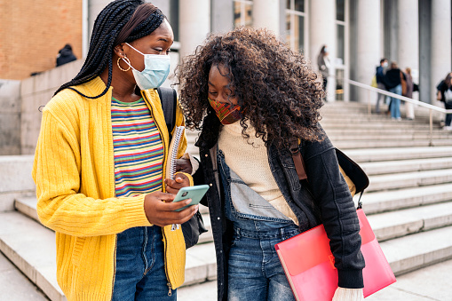 Stock photo of black female students talking and using phone. They are wearing face mask due to covid19.