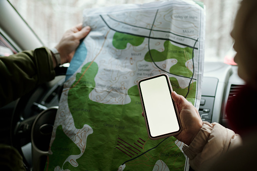 Close-up of hand of mature woman holding smartphone over local paper map held by her husband during their travel by car in winter