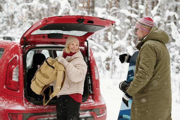 Adult man in winter clothing with knitted hat puts something into white car trunk in snowy day. Winter season concept.