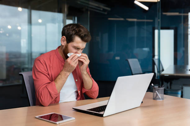 istock Sick man at work has flu and cold, businessman sneezes and coughs at workplace working inside office at desk using laptop at work 1465813598