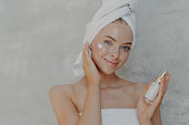 Beautiful tender smiling woman applies face cream, takes care of her beauty, has natural makeup, clean perfect skin, wrapped in bath towel, poses against grey background. Beauty treatments concept