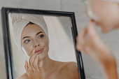 Healthy woman wears minimal makeup, takes care of complexion and lips, looks at herself in mirror, stands bare shoulders, wears bath towel on head, has healthy flawless skin. Beauty, hygiene concept