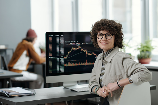 Young smiling businesswoman or crypto analyst in casualwear sitting by workplace with computer monitor and graphic data on screen