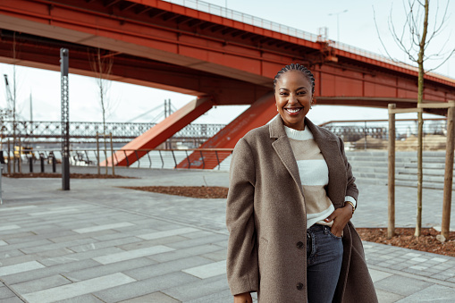 A beautiful African-American woman in casual clothing is smiling, looking at the camera, and posing outdoors on a sunny Winter day. There is a red bridge behind her.