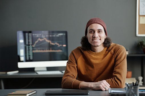 Young smiling male broker in casualwear looking at camera while sitting by workplace against computer screen with financial graphic data