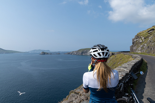A shot of a young woman in a helmet with her bike looking at the ocean at Slea Head, Coumeenoole, Ireland