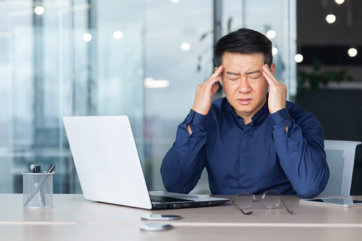 Tired young man Asian businessman feels severe headache migraine. He sits at his desk holds his head with his hands closes his eyes
