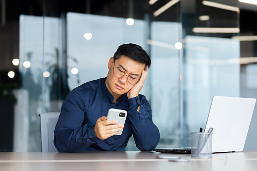 Portrait of a worried young Asian man sitting in the office at the table, holding his head with his hand, looking sadly at the phone. Bored, received bad news, messages.
