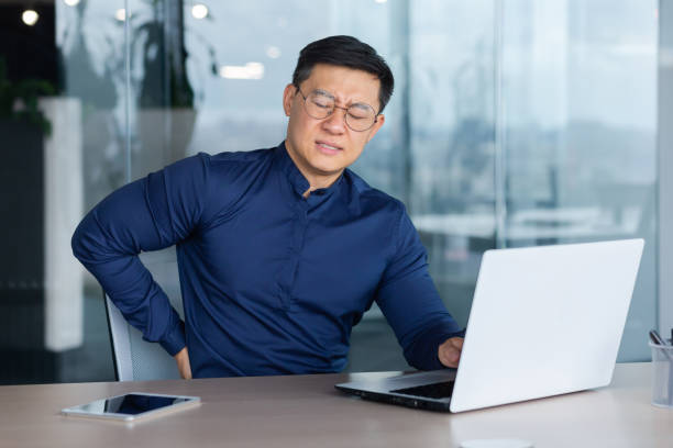 ein junger asiatischer student sitzt mit einem laptop im büro. er spürt starke schmerzen im rücken - back rear view men muscular build stock-fotos und bilder