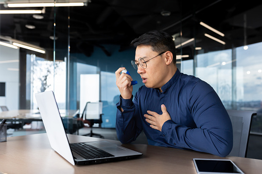 Young male Asian office worker having asthma attack at workplace, breathing hard. He sits at a table with a laptop, uses an inhaler.