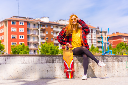 A stylish redhead Hispanic lady enjoying a sunny day in an urban park holding her skateboard