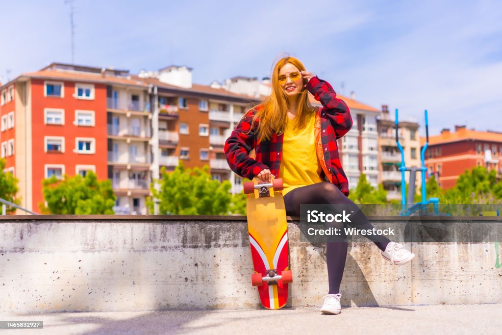 Stylish redhead Hispanic lady enjoying a sunny day in an urban park holding her skateboard A stylish redhead Hispanic lady enjoying a sunny day in an urban park holding her skateboard Hipster Culture Stock Photo