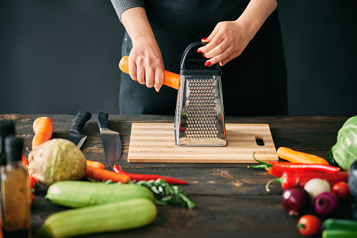 Woman cooking in kitchen at home. Cropped image of female hands rubs carrots on a grater. Culinary, healthy eating concept