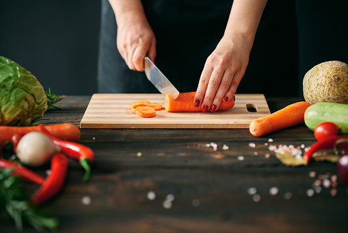 Woman cooking in kitchen at home. Closeup of female hands slicing carrots on chopping board. Culinary, healthy eating concept