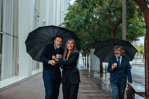 Three mature businesspeople are walking in the city street in the rain with coffee cups on a rainy day.