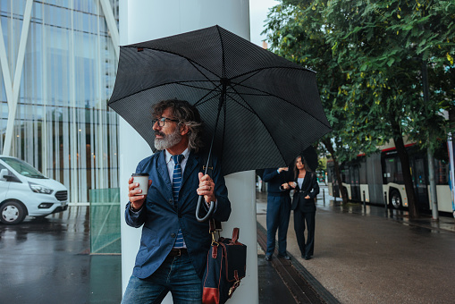 A business executive is standing outside with an umbrella and a coffee to go in the street.