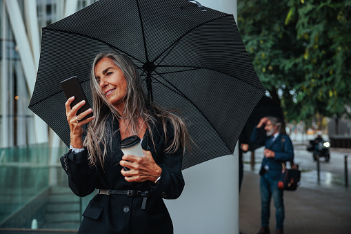 A smiling Caucasian businesswoman with a telephone and an umbrella is outside on a rainy day.