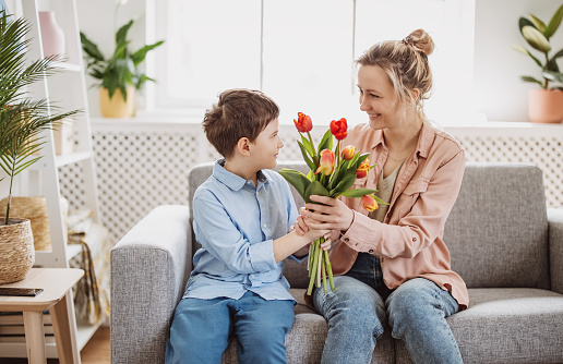 Cute boy sitting on the sofa with mom and giving a bouquet of tulips to her. Concept of the Mother's day and family relation.