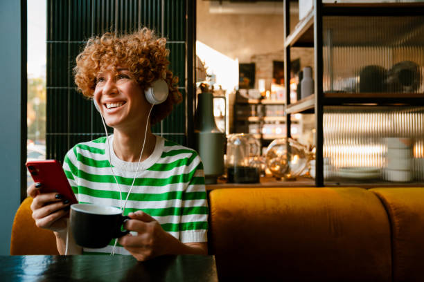 Jeune femme gingembre bouclée dans des écouteurs utilisant un téléphone portable tout en buvant du café - Photo