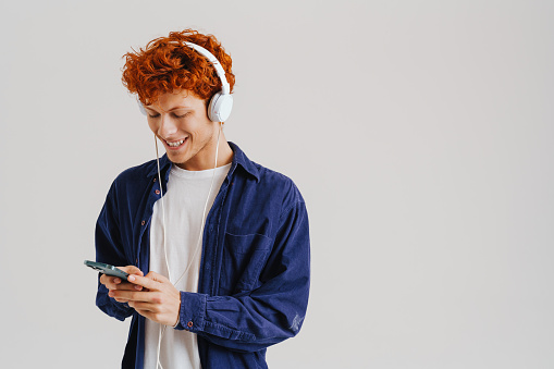 Ginger man in headphones using cellphone while listening music isolated over white background