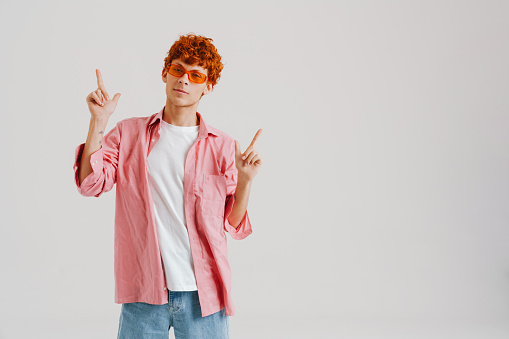 Young ginger man wearing sunglasses pointing fingers upwards isolated over white background