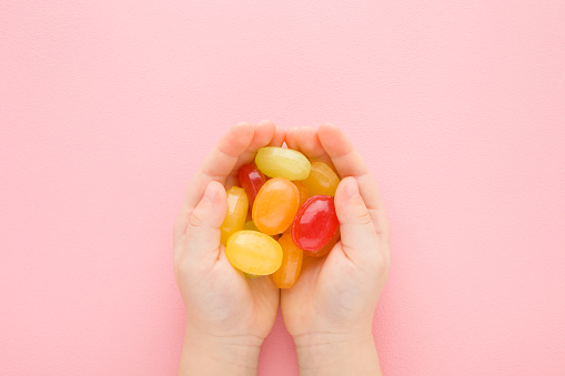 Colorful sugar candies in baby girl opened palms on light pink table background. Pastel color. Sweet snack. Closeup. Point of view shot. Top down view.
