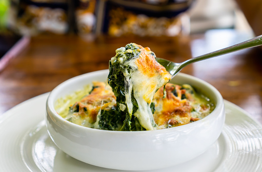 Spoon scoop up baked Spinach with Cheese in white bowl.