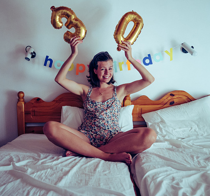 Woman celebrating her birthday. Holding a large inflatable 30 sign balloon. Morning surprise in bed