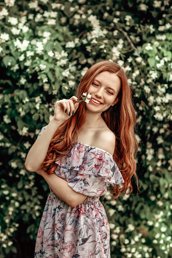 A girl in a light summer dress stands near a bird cherry bush on a hot summer day. She is holding white flowers in her hands. The girl smiles