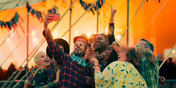 A medium group of friends at a festival all dancing together and having a good time outside an entertainment tent.. Everyone is happy and smiling and sharing positive energy. They are having a group selfie taken together on a friends orange smartphone. Bright orange lights light up the inside of the Large tent.