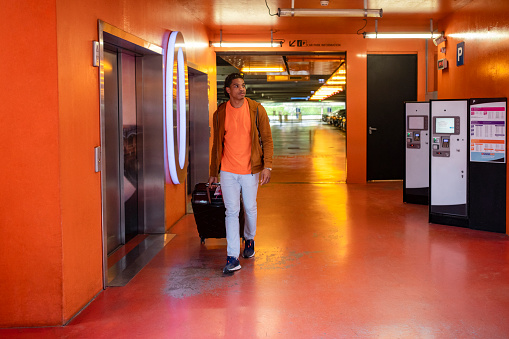 A young man walking through a multi-storey car park at an airport in Toulouse, France. He is walking past the pay area while pulling his suitcase behind him.