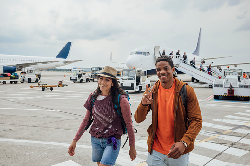 A young woman and man walking outdoors on a zebra crossing on an airport tarmac in Toulouse, France after getting off their flight. The man is doing the peace sign and looking at the camera as they both smile.