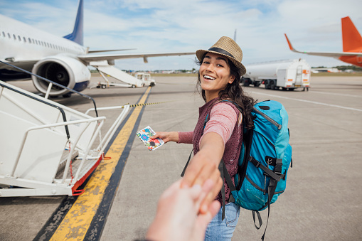 A personal perspective of a young woman walking outdoors on an airport tarmac in Toulouse, France while looking over her shoulder at the camera and smiling. She is holding hands with an unrecognisable person and leading them to the plane.