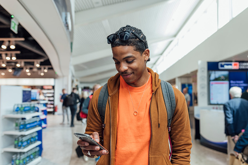 A young man walking through an airport in Toulouse, France while text messaging a friend on his mobile phone and smiling.