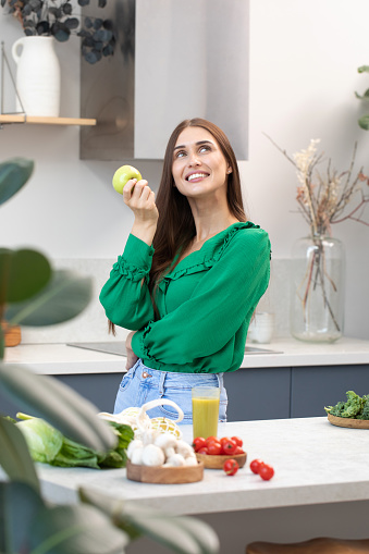 A happy young woman holds a green apple in her hand in the kitchen. The concept of healthy eating, vegetarianism.