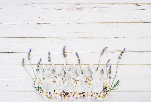 Flat lay of white and grey wooden vintage easter bunny shapes with lavender and almond blossoms on white wooden background with copy space. Color editing with added grain. Very selective and soft focus. Part of a series.
