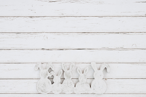 Flat lay of white and grey wooden easter bunny shapes on white wooden background with generous copy space. Color editing with added grain. Very selective and soft focus. Part of a series.