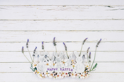 Flat lay of white and grey wooden vintage easter bunny shapes with lavender and almond blossoms and the lettering happy easter on white wooden background with copy space. Color editing with added grain. Very selective and soft focus. Part of a series.