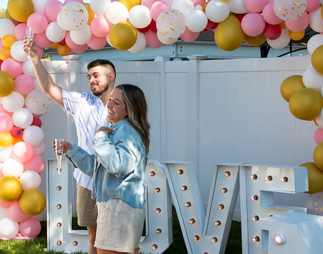Just engaged couple raising a glass of champaign to their family standing under baloons and in front of a love sign after she said yes.