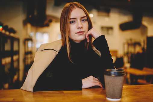 Charming young woman in stylish clothes sitting at wooden table with cup of takeaway beverage and looking at camera against cozy cafe