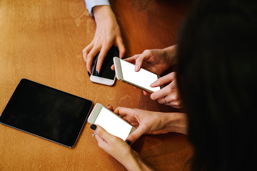 From above of crop faceless ladies text messaging via smartphones while gathering together at wooden table with modern portable tablet