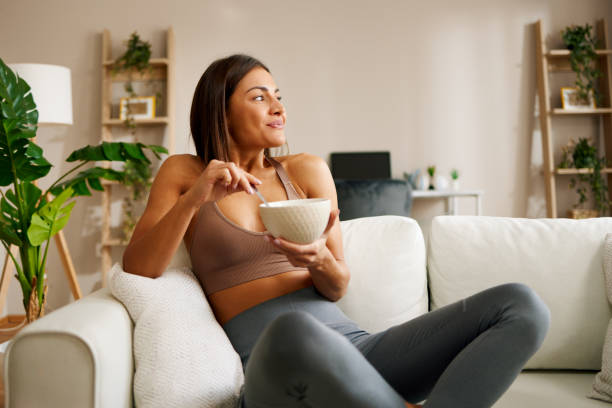 beautiful young woman is sitting on the sofa in the living room and eating oatmeal - eating women breakfast cereal imagens e fotografias de stock