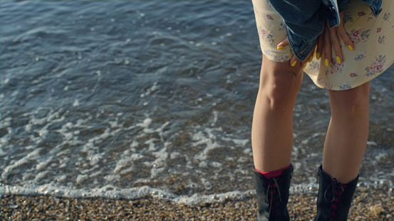 Closeup stylish girl legs standing at beach. Unrecognizable woman in casual clothes posing at seaside. Female hipster feet wearing trendy boots. Ocean waves splashing on sandy shore. Summer concept