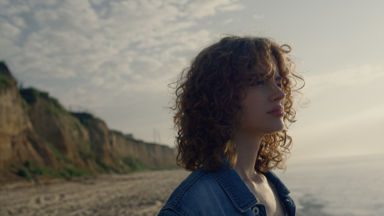 Portrait of curly woman looking around ocean landscape at sunrise. Thoughtful girl enjoying sea view at rocky coastline. Female model turning head around. Relaxed lady blinking eyes. Leisure concept