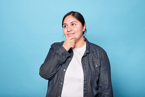 Young curvy latina woman looks empty space, hand on chin, wearing denim shirt and hoop earrings and tied hair. Indoor studio shot isolated on blue background.