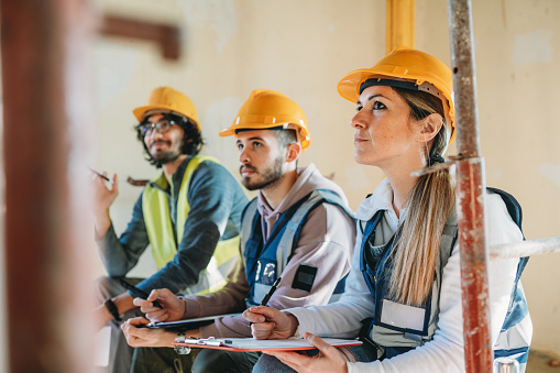 Three people are listening to their foreman on a construction site. They are sitting on the worksite.