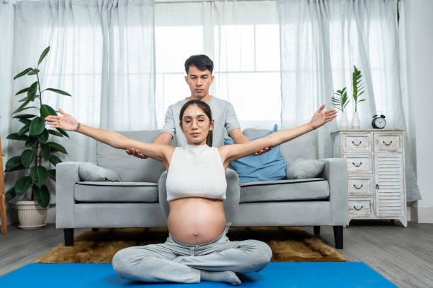 경찰서 거실의 요가 매트에 앉아 있는 아시아 남성과 여성. 배가 큰 임산부를 돕고 있는 아버지 출산을 앞둔 며칠 동안 휴식을 취하기 위해 요가 운동을 해보세요. - torso physical therapy patient relaxation exercise 뉴스 사진 이미지