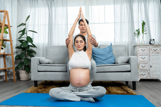 uomo e donna asiatici siedono su un tappetino da yoga nel soggiorno di una casa di polizia. un padre che sta aiutando una madre incinta con una grande pancia prova lo yoga per rilassarti nei giorni prima del parto. - torso physical therapy patient relaxation exercise foto e immagini stock